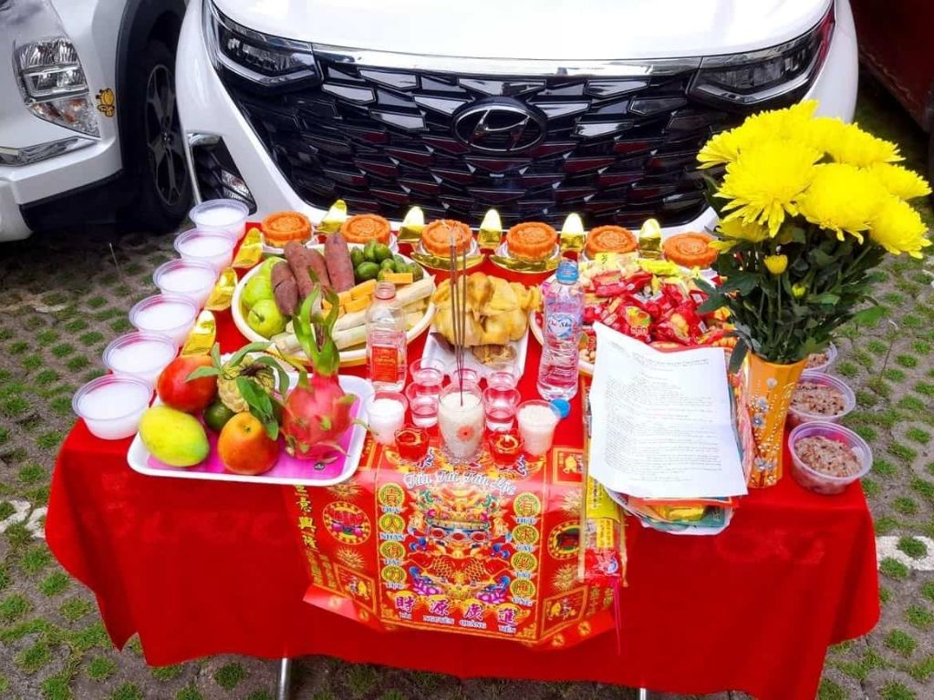Mâm cúng xe mới đầy đủ lễ vật gồm trái cây, hoa, bánh kẹo và vàng mã đặt trước đầu xe ô tô mới mua.