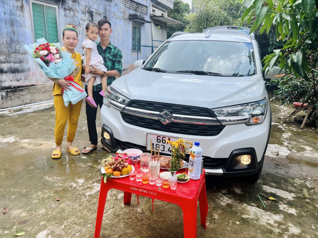 Gia đình tổ chức lễ cúng xe mới tại nhà, bàn cúng bày lễ vật trước đầu xe Suzuki trắng.
